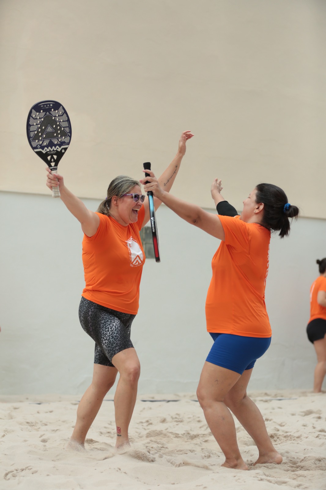 Duas jogadoras de camiseta laranja comemoram com os braços e as raquetes erguidos na quadra de areia da Arena Aldeia